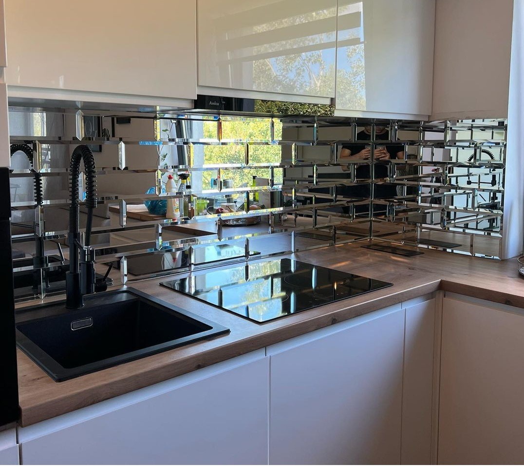 Kuchnia z lustrzaną mozaiką na ścianie nad blatem, czarny zlew i bateria, płyta indukcyjna, białe szafki, drewniany blat.