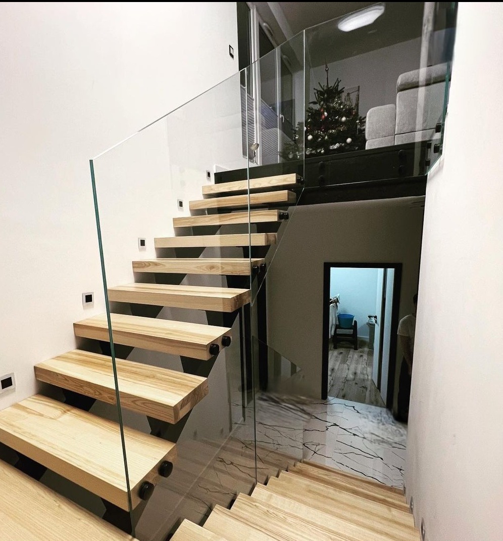 Nowoczesne schody z drewnianymi stopniami i szklaną balustradą, widok z dołu, w tle salon z choinką.