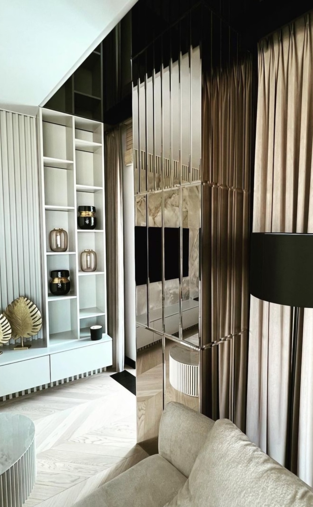 Nowoczesny salon z podziałem przestrzeni za pomocą pionowych paneli lustrzanych, widoczny regał z dekoracjami i beżowe zasłony.
