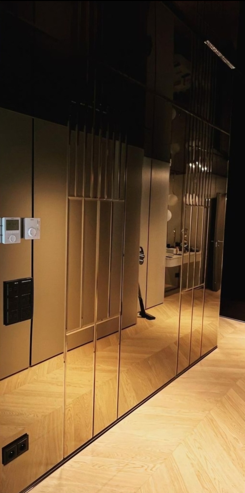 Wnętrze z lustrzaną ścianą z pionowymi podziałami, odbijającą fragment pokoju z parkietem w jodełkę i elementami wyposażenia.