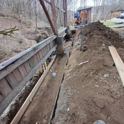 Bartek-Bruk - Budowa mostku nad ciekiem wodnym: wykop z szalunkiem, w tle koparka i sterta ziemi. Widoczne elementy drewnianej konstrukcji i fragment buta pracownika.