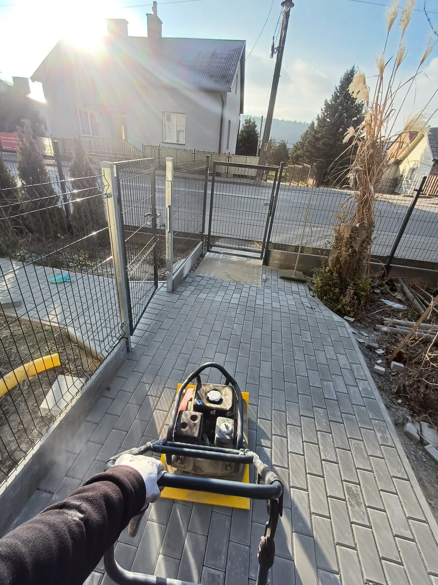 Zagęszczarka w dłoniach pracownika ubija świeżo ułożoną, szarą kostkę brukową o prostokątnym kształcie na podjeździe przed domem z metalowym ogrodzeniem w słoneczny dzień.