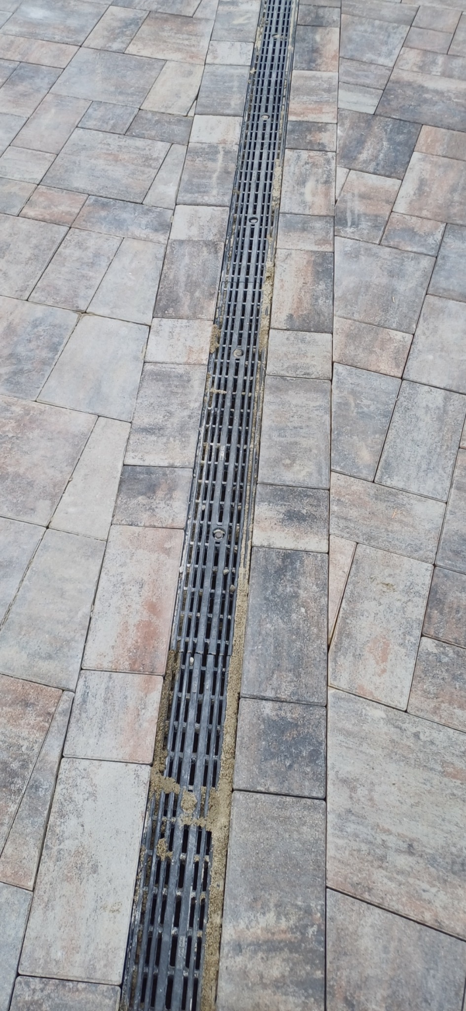 Ułożona kostka brukowa w odcieniach szarości i beżu z zamontowanym liniowym odpływem wody, wypełnionym piaskiem.