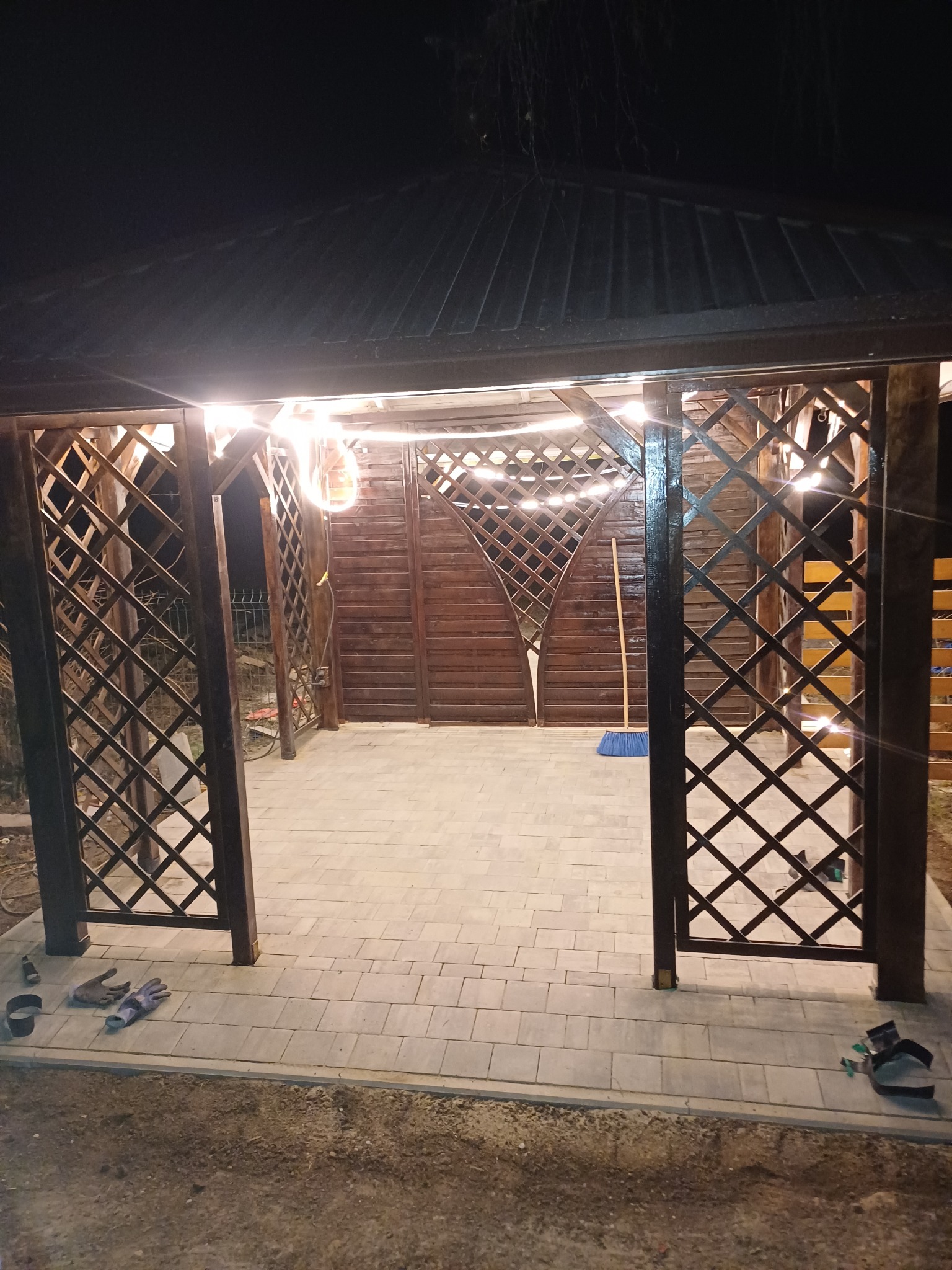 Zadaszona altana z drewnianą kratą i oświetleniem sznurkowym, wyłożona jasną kostką brukową, widoczne rękawice robocze i szczotka.