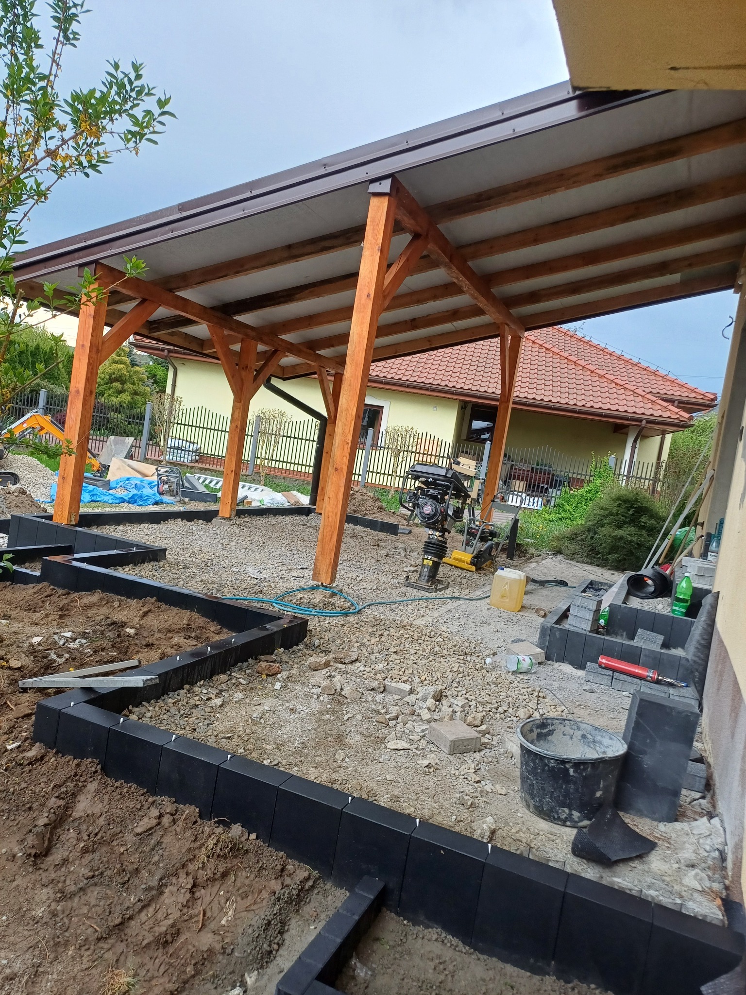 Etap budowy zadaszenia i obrzeży rabat z kostki. Widoczna ubijarka, narzędzia i materiały budowlane. W tle dom z czerwoną dachówką.