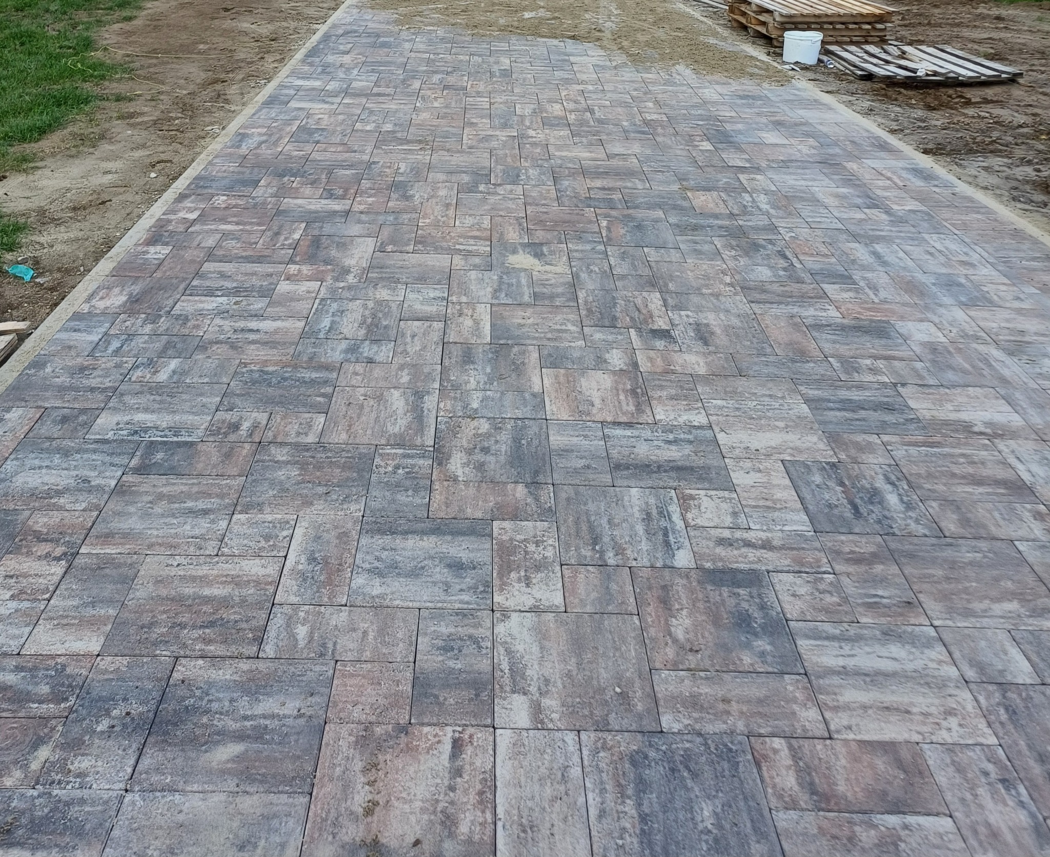 Ukończona ścieżka z kostki brukowej w odcieniach szarości i brązu, ułożona we wzór. Widoczne palety w tle, co sugeruje świeżo zakończone prace.