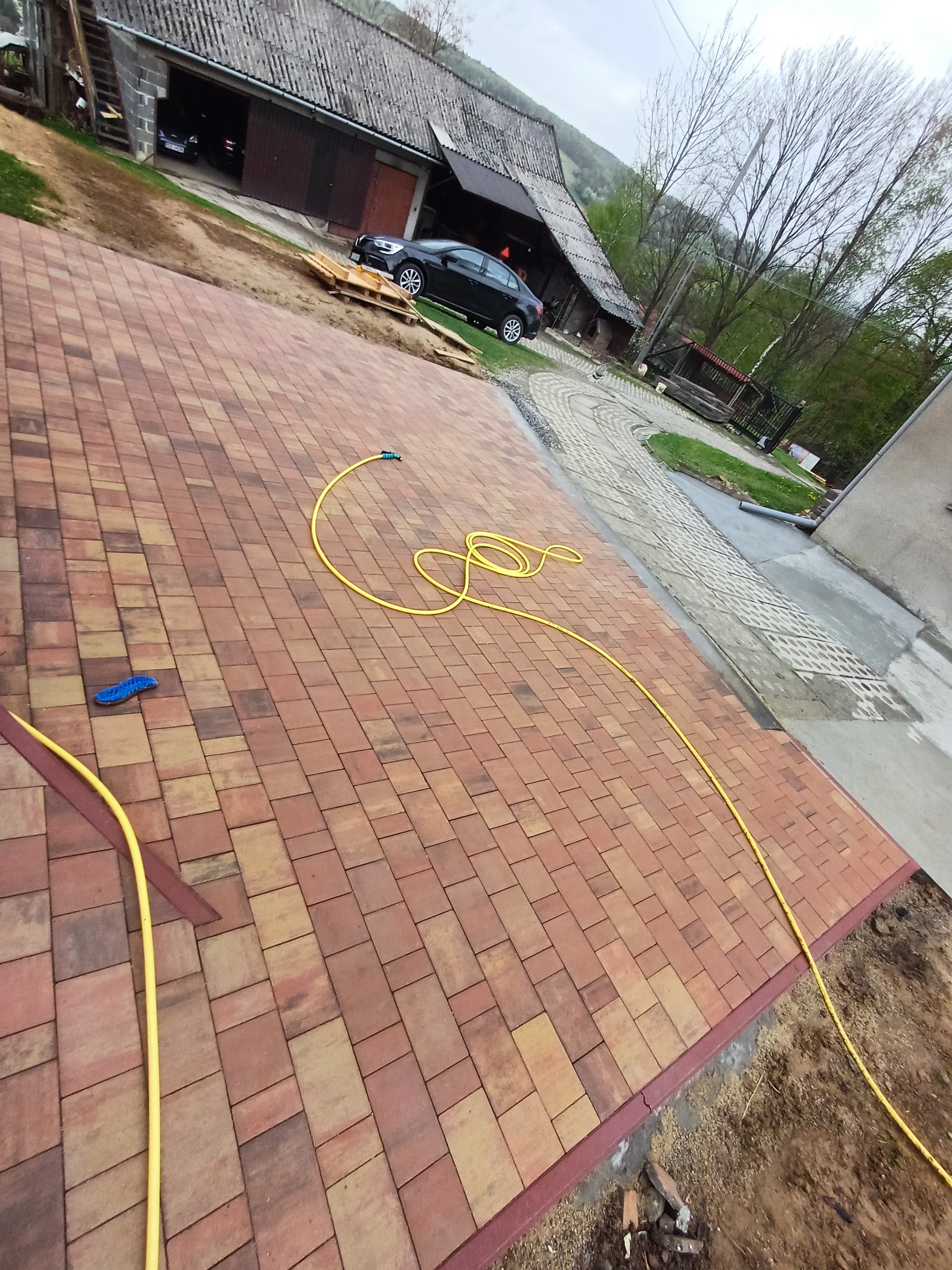 Ukończona nawierzchnia z kostki brukowej w odcieniach czerwieni i brązu. Żółty wąż ogrodowy leży na kostce. W tle budynek gospodarczy i drzewa.