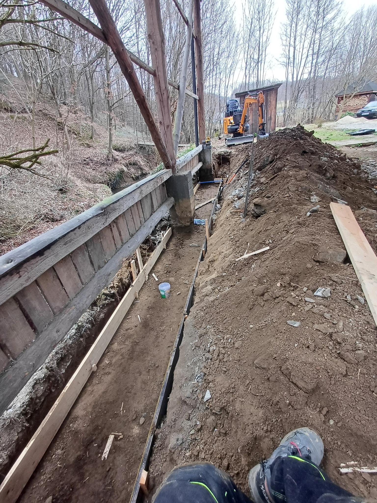 Budowa mostku nad ciekiem wodnym: wykop z szalunkiem, w tle koparka i sterta ziemi. Widoczne elementy drewnianej konstrukcji i fragment buta pracownika.