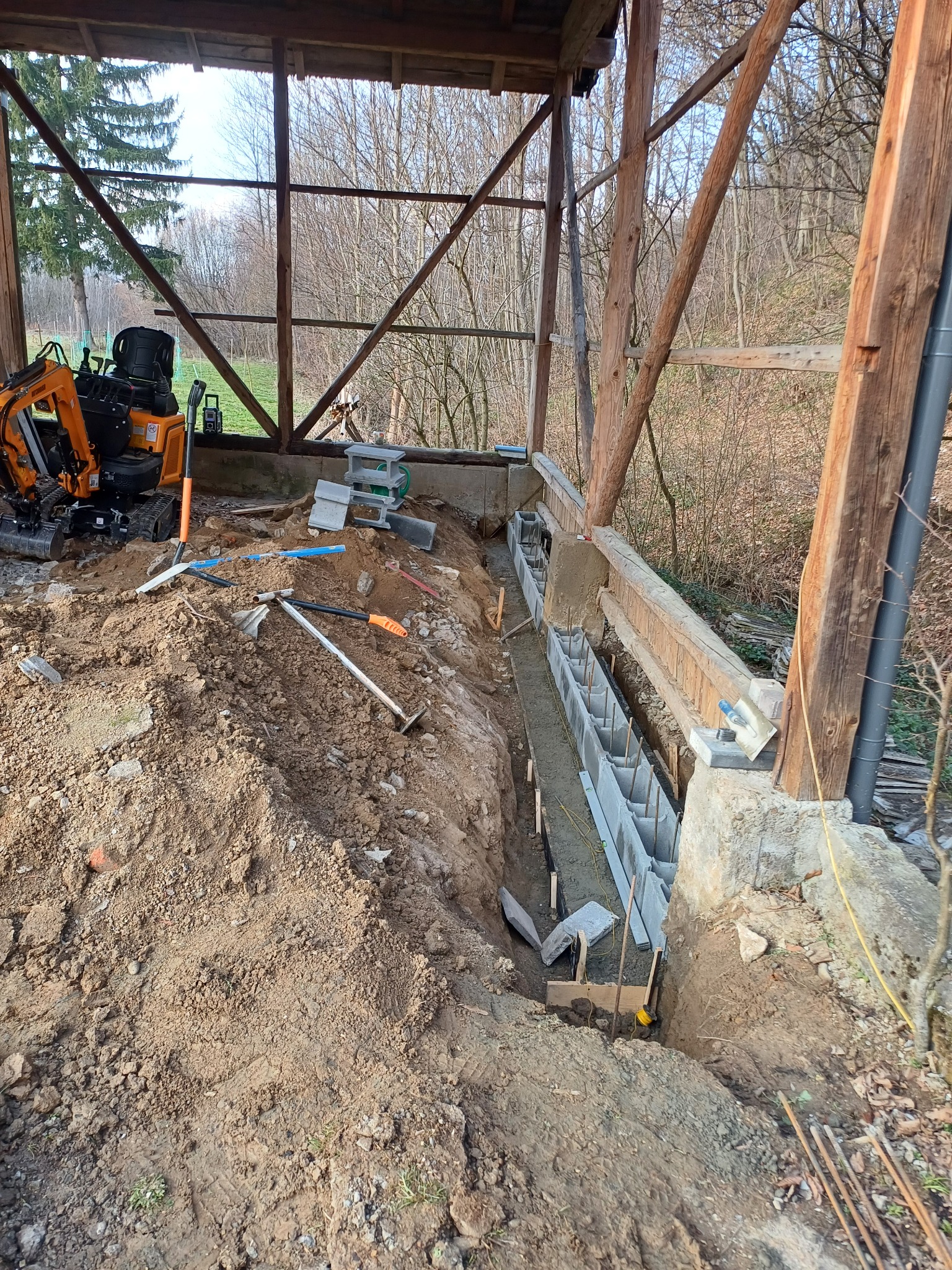 Fundamenty pod budynek w trakcie budowy, widoczny wykop, szalunki z betonu komórkowego oraz mini koparka na ziemi.