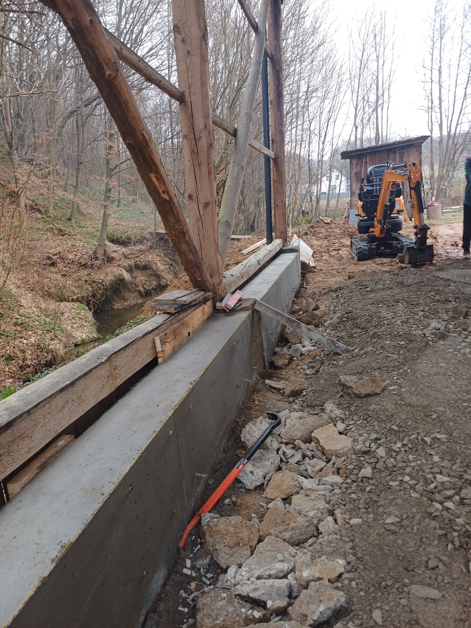Betonowa podpora mostu w trakcie budowy z drewnianą konstrukcją, widoczny mini-koparka i potok w tle. Prace ziemne w toku.