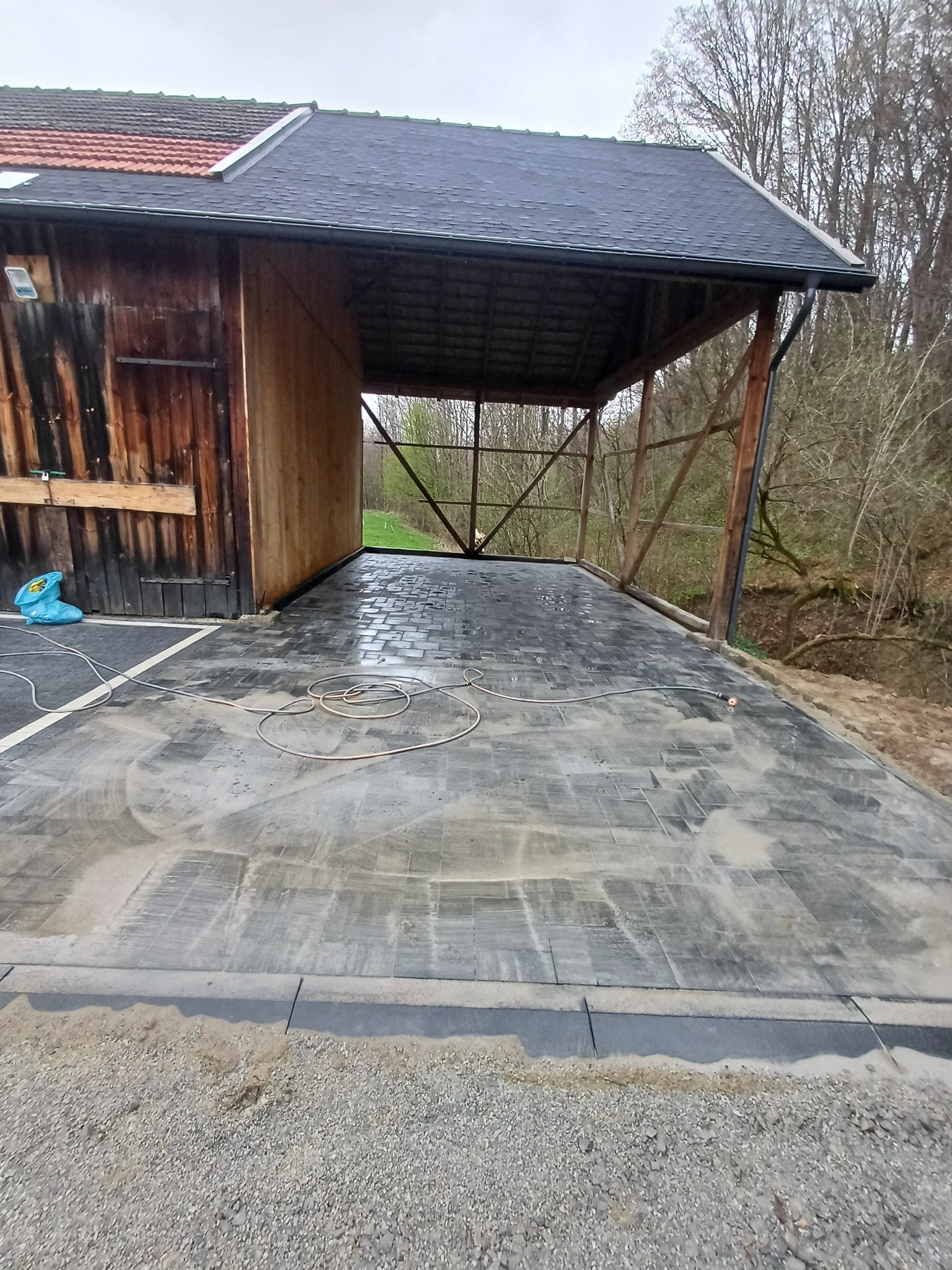 Zadaszona wiata z ułożoną, szarą kostką brukową. Widoczne kable i niebieska torba. W tle drzewa i fragment lasu. Kąt ujęcia z dołu, lekko z prawej strony.