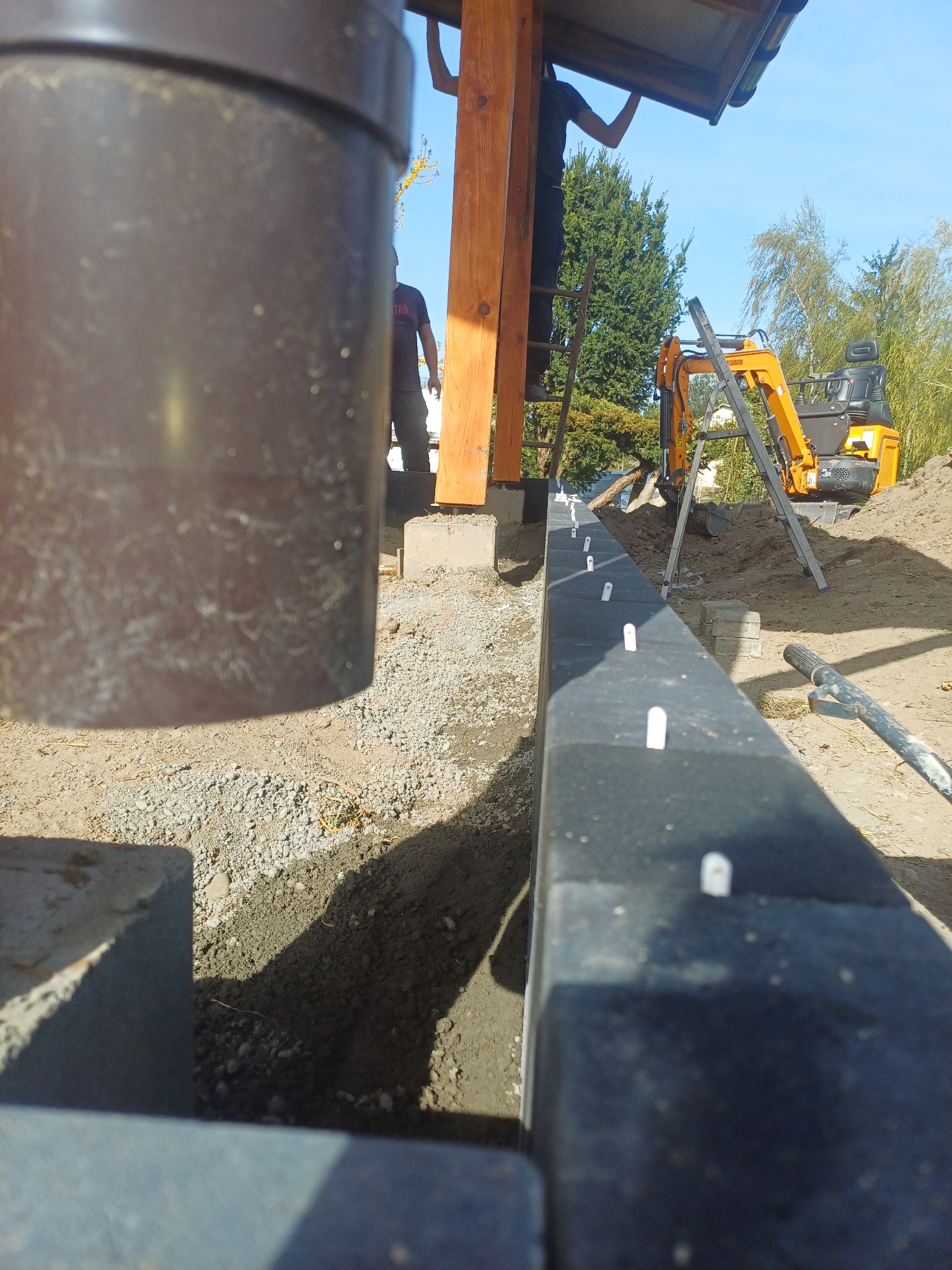 Przygotowanie terenu pod budowę, widoczny wykop, obrzeża z kołkami stabilizującymi, mini koparka w tle i pracownicy na drabinie.