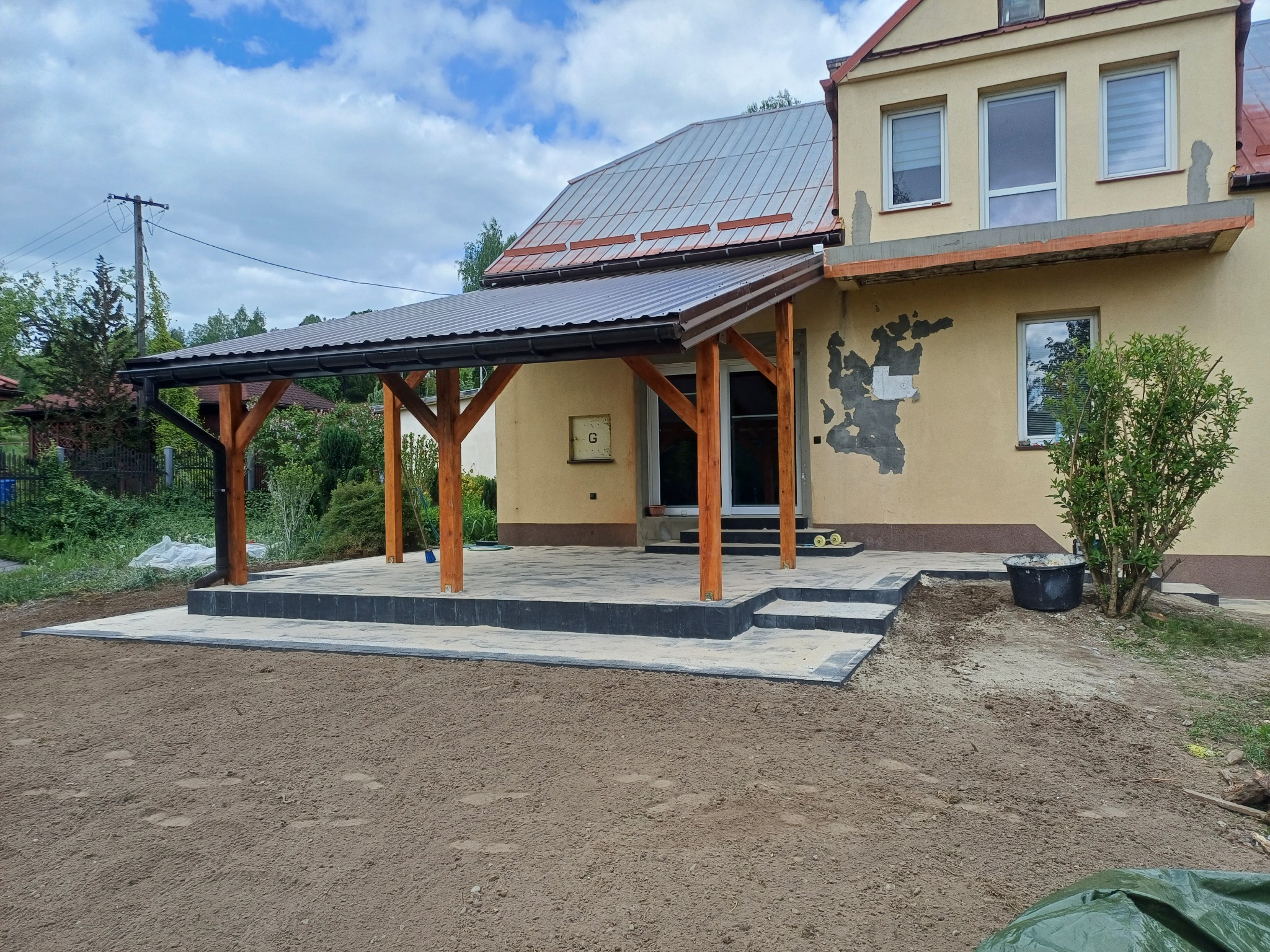 Zadaszony taras z kostki brukowej w odcieniach szarości, z drewnianymi słupami i schodami, otoczony przygotowanym podłożem gruntowym. Widok na budynek w tle.