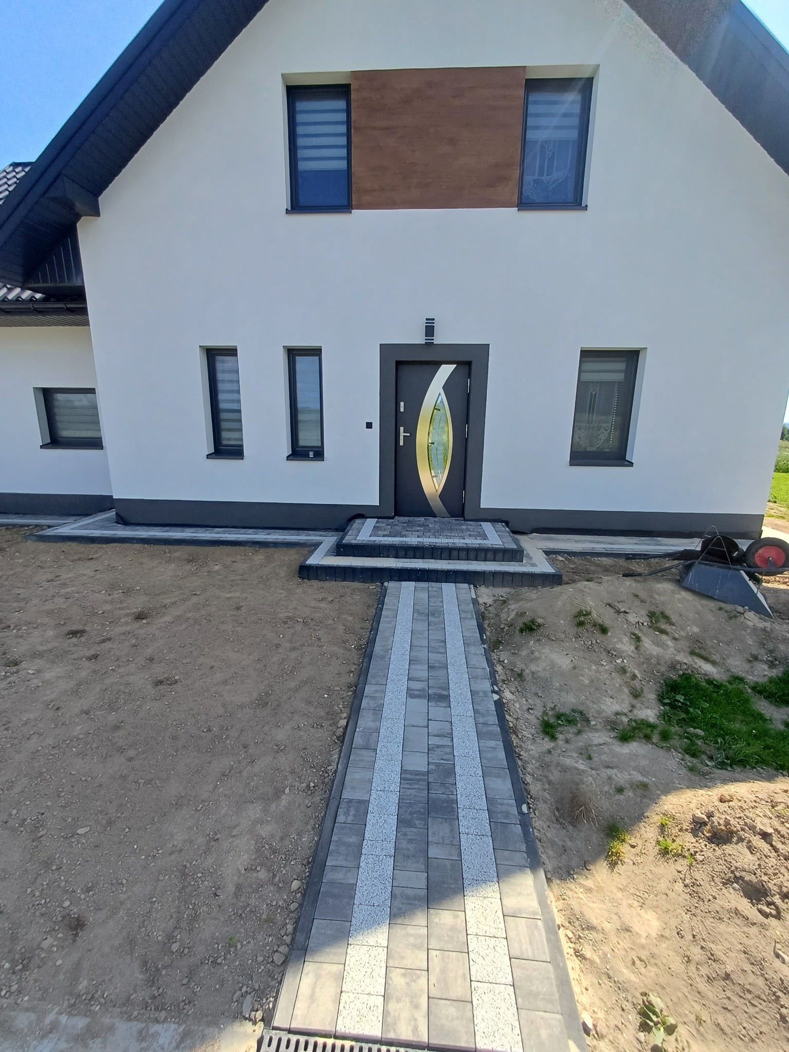 Nowoczesny dom z szarą kostką brukową tworzącą ścieżkę do wejścia. Uwagę zwraca symetryczny układ kostki i kontrast z jasną elewacją.