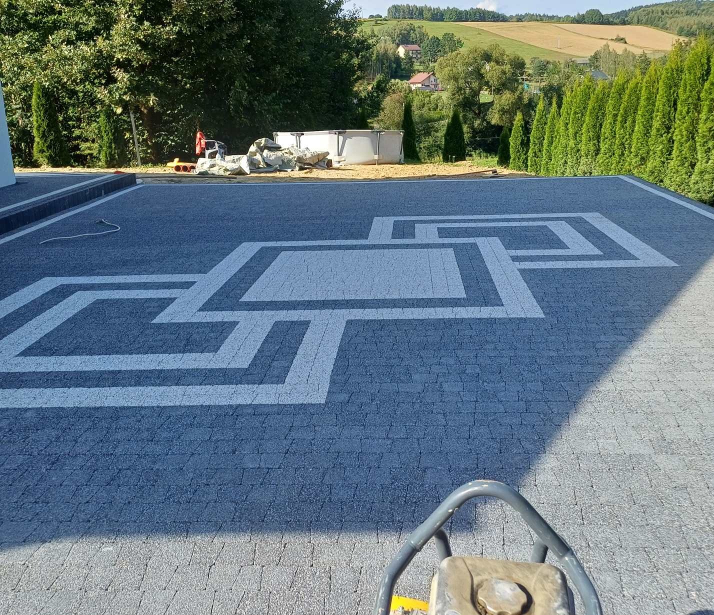 Ułożona kostka brukowa z geometrycznym wzorem w Jodłowej, widok z góry z wibratorem do zagęszczania. W tle zielony krajobraz.