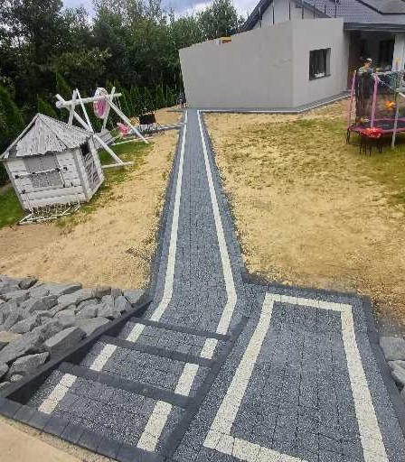Ukończona ścieżka z kostki brukowej w ogrodzie, z białymi akcentami, prowadząca od schodów do placu zabaw i budynku. Widok z góry na szare kostki.