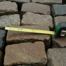 Pomiar długości kamienia brukowego typu chlebówka za pomocą zwijanej miarki, widoczne różne odcienie szarości i brązu na powierzchni kamieni.