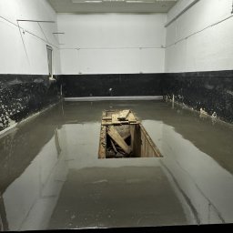 Artur Posadzki Przemysłowe - Świeżo wylana, mokra posadzka w garażu w Przeworsku, z widocznym otworem inspekcyjnym zabezpieczonym drewnianymi deskami. Ściany pomalowane do połowy na czarno, górna część biała.