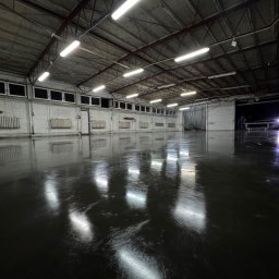 Artur Posadzki Przemysłowe - Błyszcząca, ciemna posadzka w przestronnej hali magazynowej z betonowym stropem i rzędami okien pod sufitem, odbijająca światło lamp.