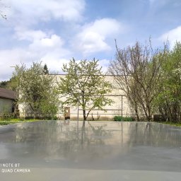 Artur Posadzki Przemysłowe - Świeżo wylana, gładka, szara powierzchnia w otoczeniu kwitnących drzew i budynku w tle, odbijająca niebo z chmurami.