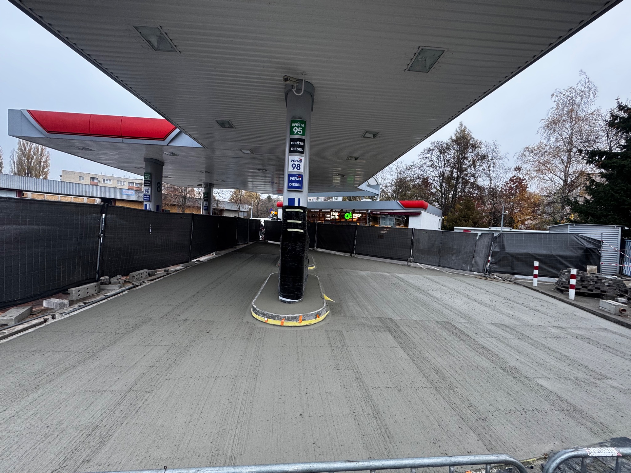 Świeżo wylana, szara wylewka betonowa pod zadaszeniem stacji benzynowej z widocznymi śladami po zacieraniu, otoczona czarną siatką zabezpieczającą teren budowy.