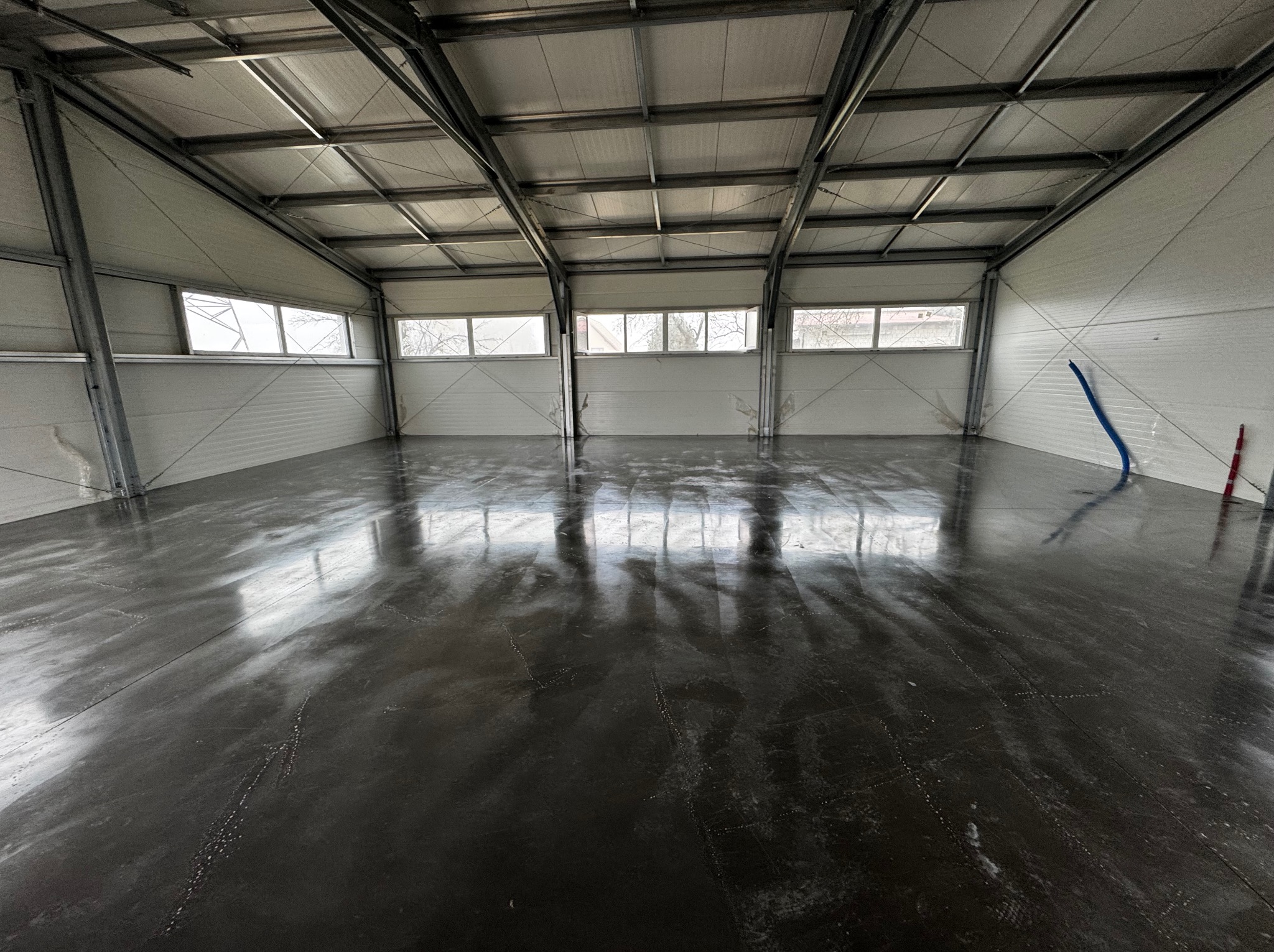 Błyszcząca, ciemnoszara posadzka w hali przemysłowej z widocznymi liniami podziału, metalowa konstrukcja dachu i białe ściany z oknami w górnej części.