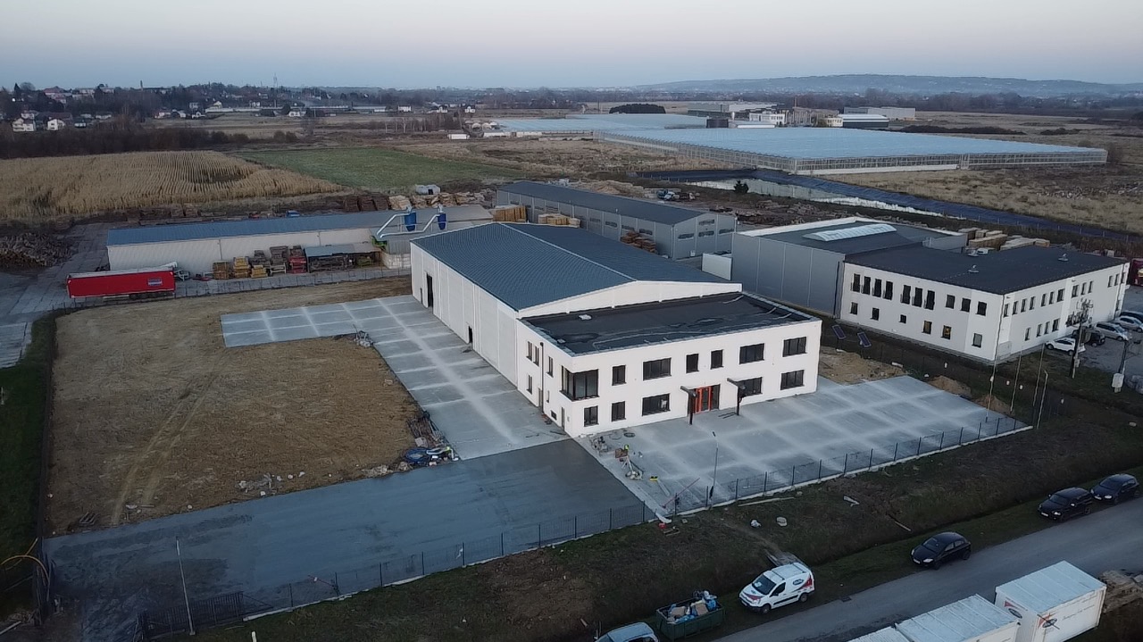 Widok z góry na nowo wylany, szary plac betonowy przed budynkiem przemysłowym, z widocznymi śladami po maszynach i świeżym betonem, w tle zabudowania gospodarcze i pola uprawne.