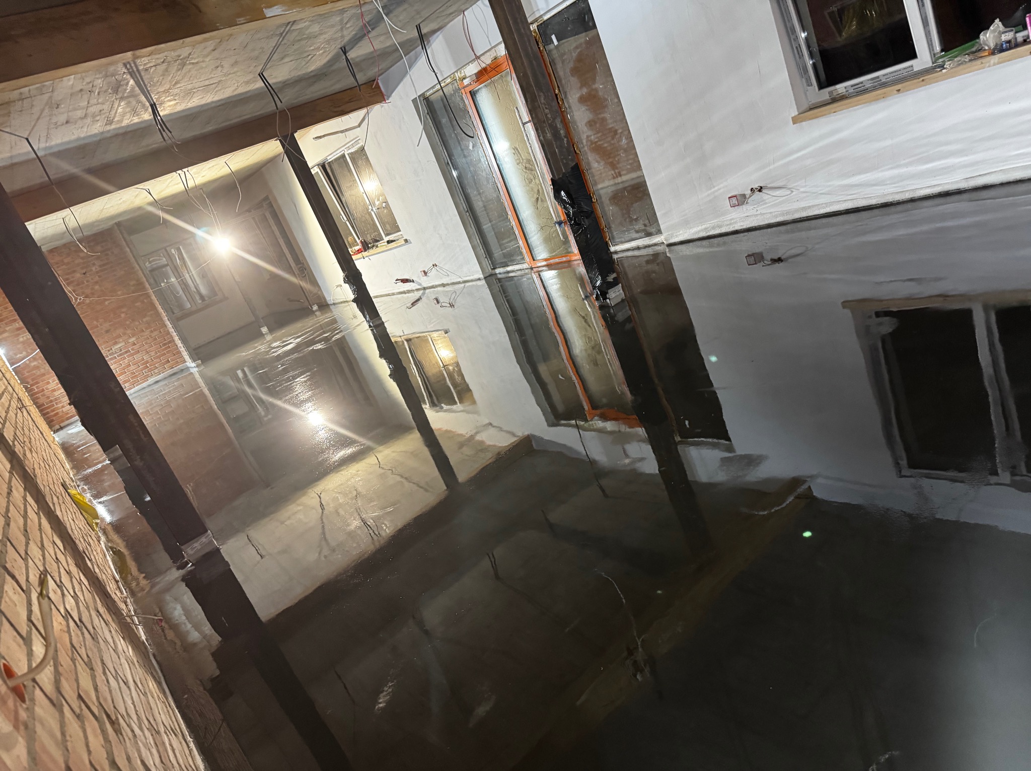 Świeżo wylana, ciemna posadzka w surowym wnętrzu z widocznymi instalacjami elektrycznymi pod sufitem i oknami w ścianach.