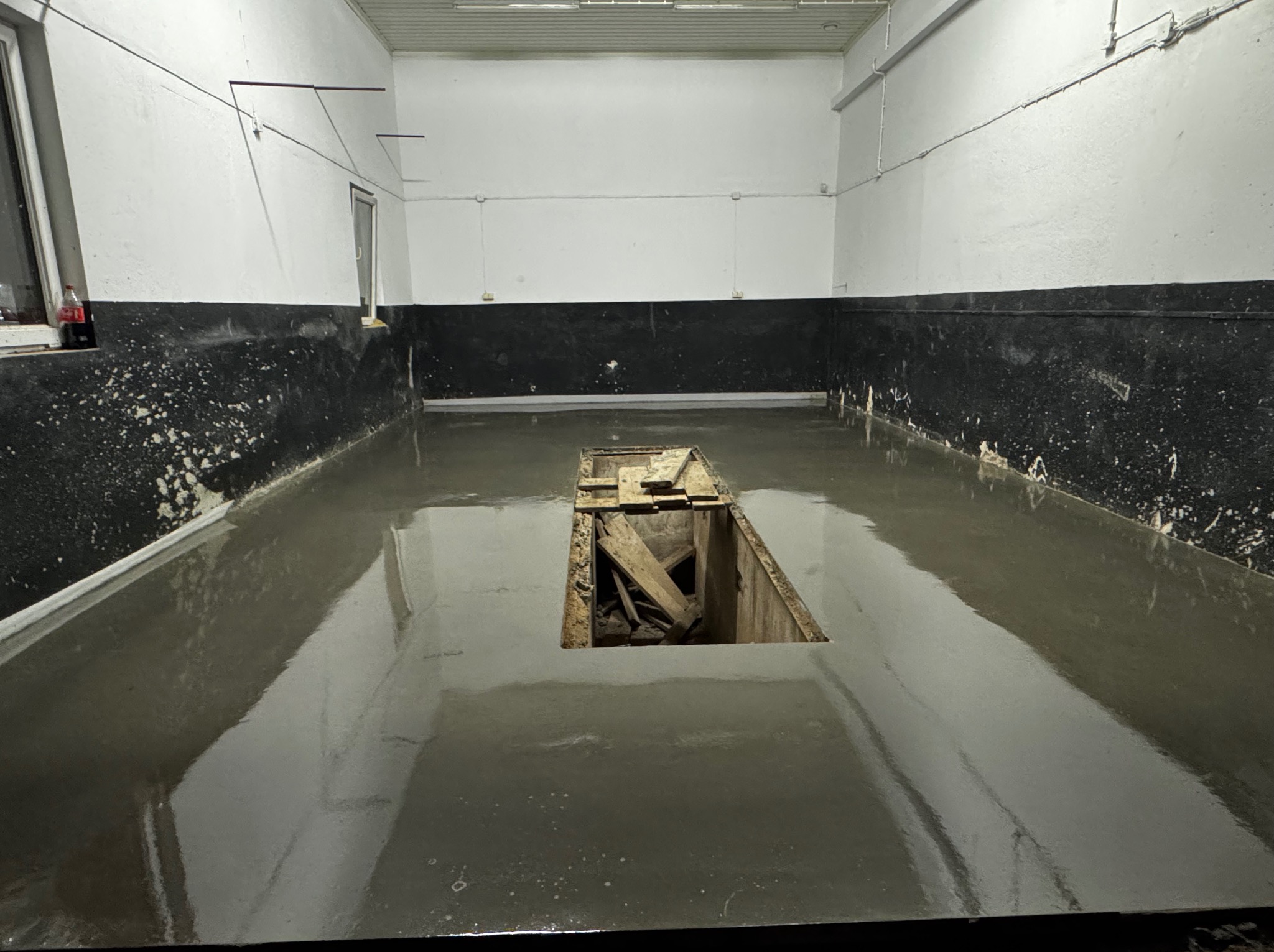 Świeżo wylana, mokra posadzka w garażu w Przeworsku, z widocznym otworem inspekcyjnym zabezpieczonym drewnianymi deskami. Ściany pomalowane do połowy na czarno, górna część biała.