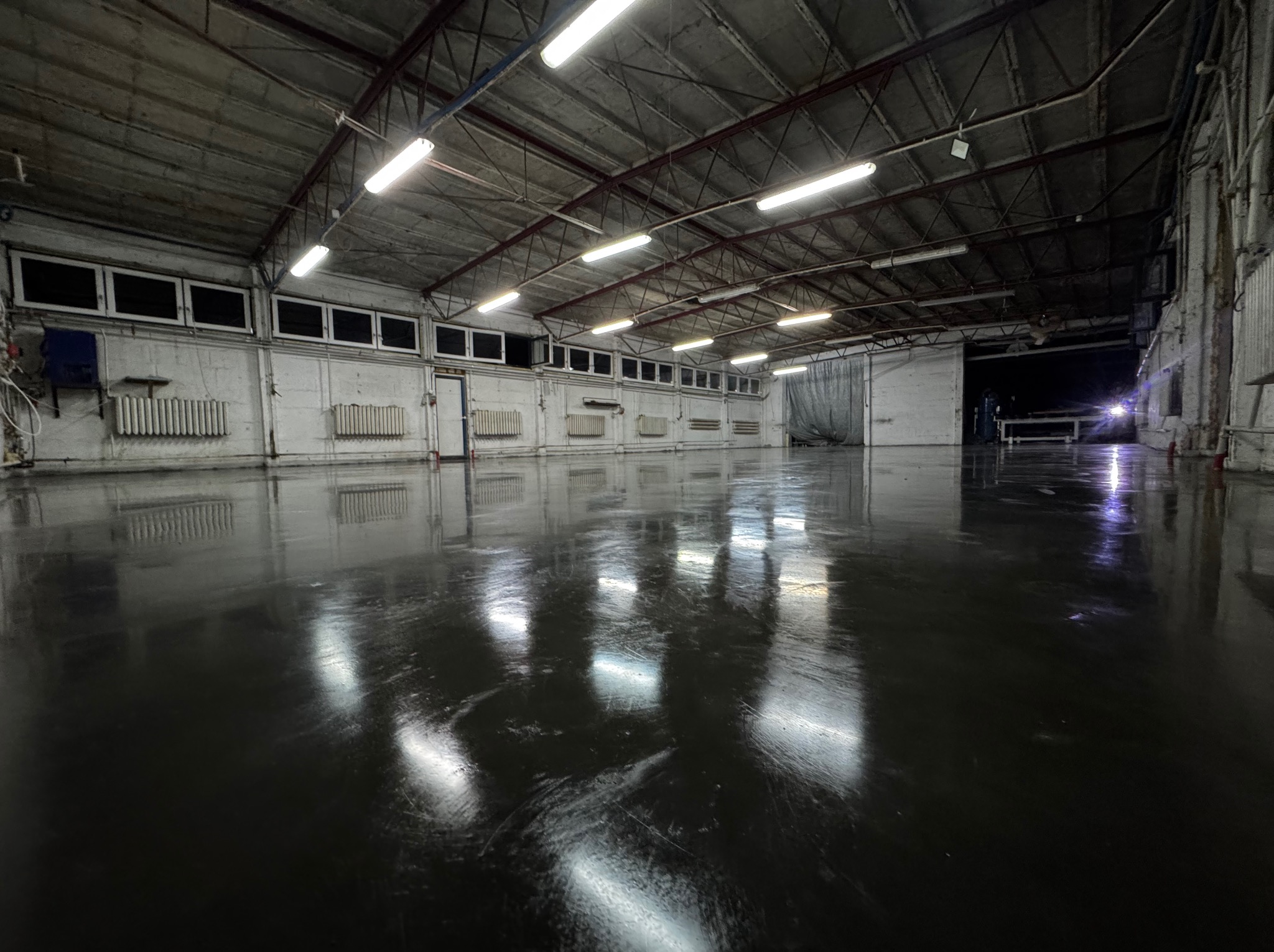 Błyszcząca, ciemna posadzka w przestronnej hali magazynowej z betonowym stropem i rzędami okien pod sufitem, odbijająca światło lamp.