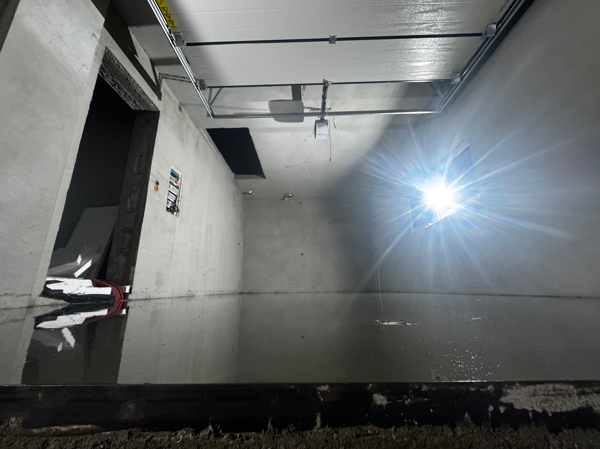 Świeżo wylana, mokra posadzka przemysłowa w surowym wnętrzu, z widocznymi elementami bramy garażowej pod sufitem i odbiciem światła lampy na powierzchni.