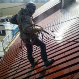 Pracownik w kasku i odzieży roboczej, zabezpieczony linami, myje metalowy dach za pomocą myjki ciśnieniowej, widoczne strumienie wody pod ciśnieniem i budynki w tle.