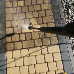 Czyszczenie kostki brukowej myjką ciśnieniową, widoczna różnica pomiędzy czystą a brudną powierzchnią, obok krawężnik i kamienie.