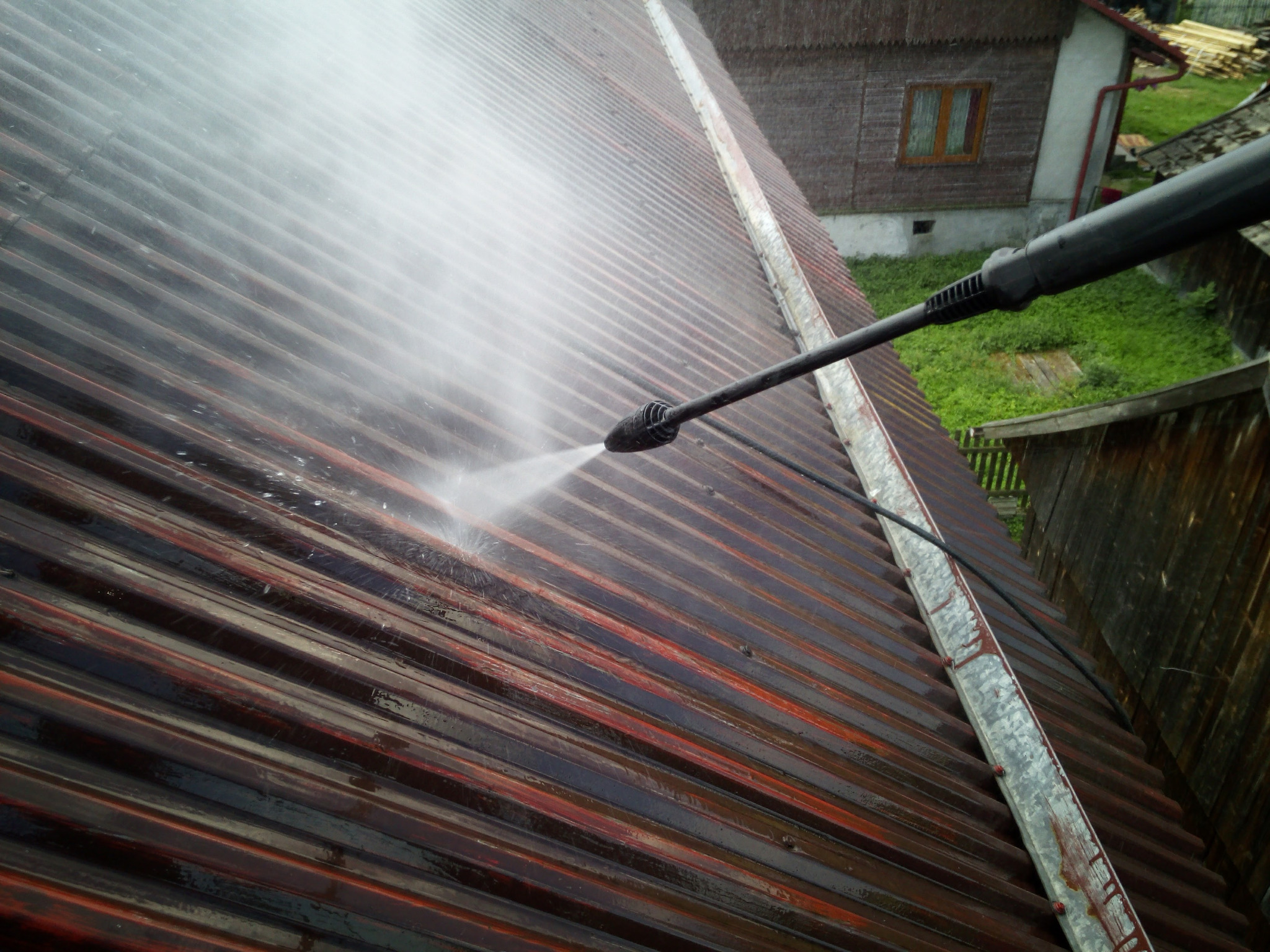 Ciemnoczerwony, blaszany dach podczas mycia pod ciśnieniem, z widocznym strumieniem wody z dyszy i fragmentem budynku w tle.