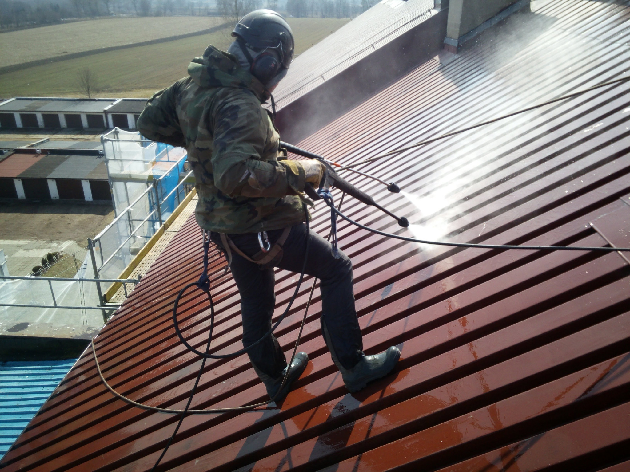 Pracownik w kasku i odzieży roboczej, zabezpieczony linami, myje metalowy dach za pomocą myjki ciśnieniowej, widoczne strumienie wody pod ciśnieniem i budynki w tle.