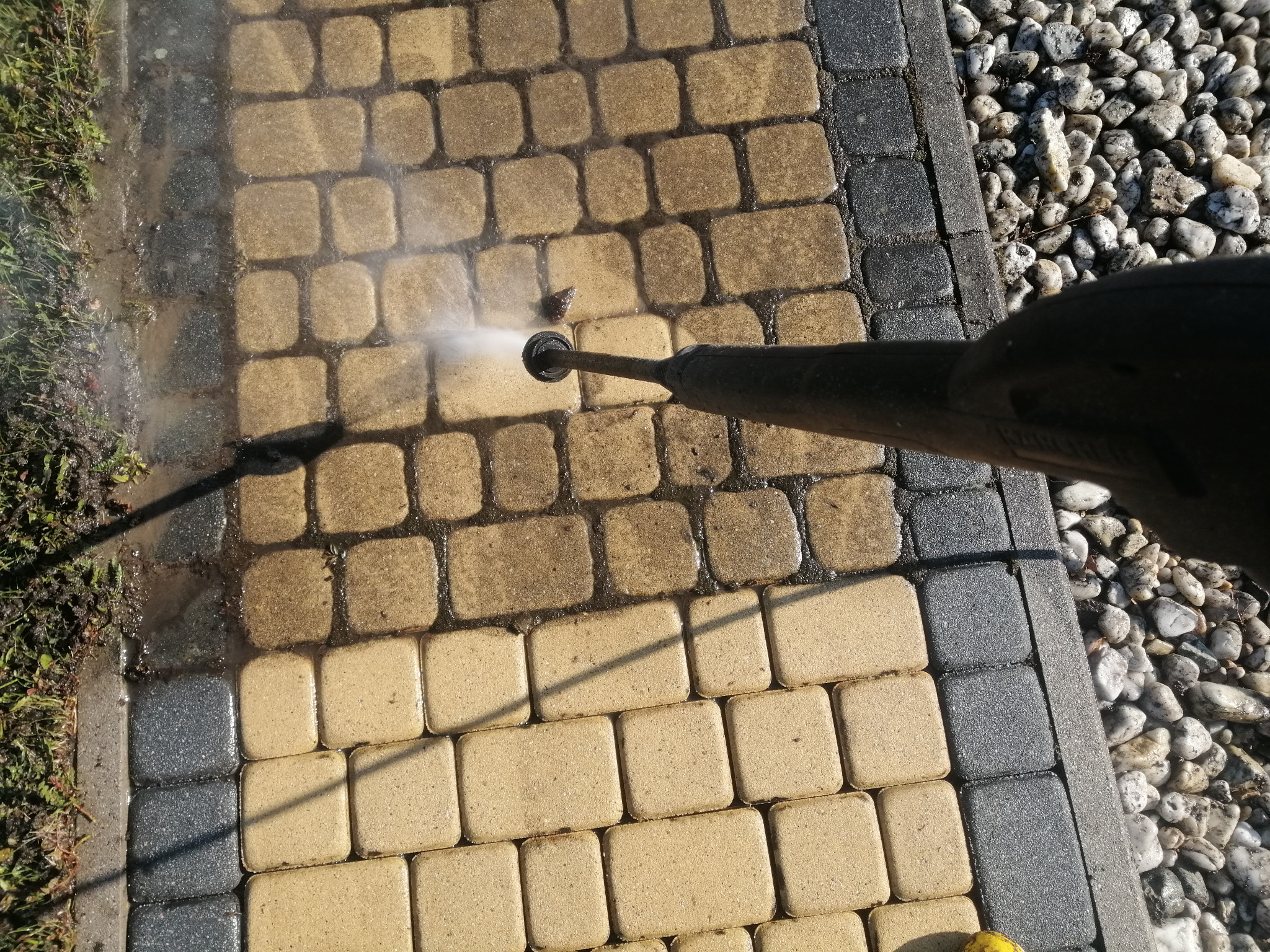 Czyszczenie kostki brukowej myjką ciśnieniową, widoczna różnica pomiędzy czystą a brudną powierzchnią, obok krawężnik i kamienie.