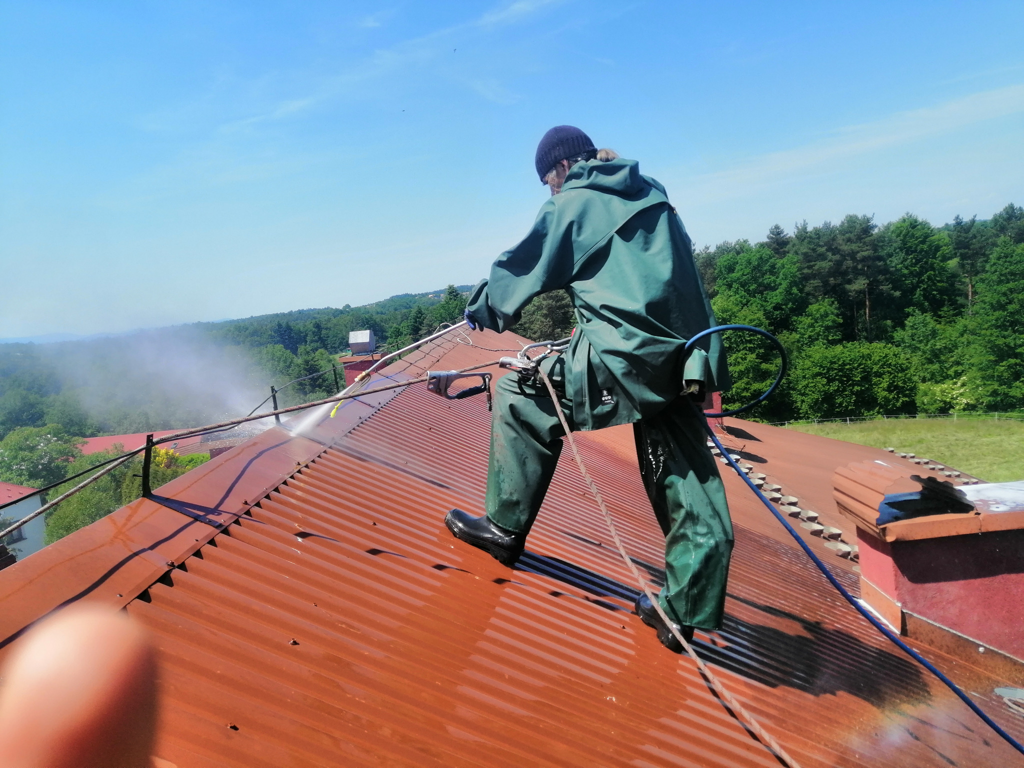 Pracownik w kombinezonie ochronnym czyści czerwoną blachodachówkę na stromym dachu za pomocą myjki ciśnieniowej, z widocznym strumieniem wody i tłem zieleni.