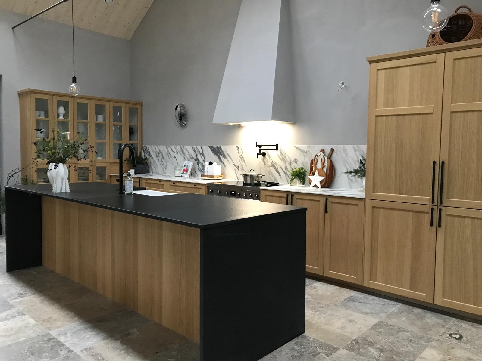 Nowoczesna kuchnia z wyspą: czarny blat, drewniane fronty szafek, marmurowe wykończenie ściany nad blatem, oświetlenie punktowe i podłużne czarne uchwyty szafek.