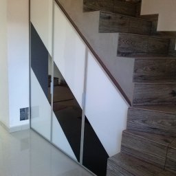 Sylwester Górski - Biało-czarna szafa wnękowa pod schodami z drewnopodobnymi płytkami, widoczna instalacja elektryczna w ścianie obok.