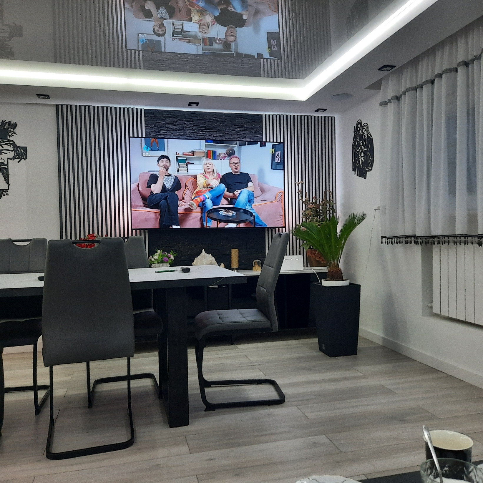 Nowoczesny salon z czarno-białą ścianą akcentującą, szare krzesła przy stole i telewizor wyświetlający program rozrywkowy.