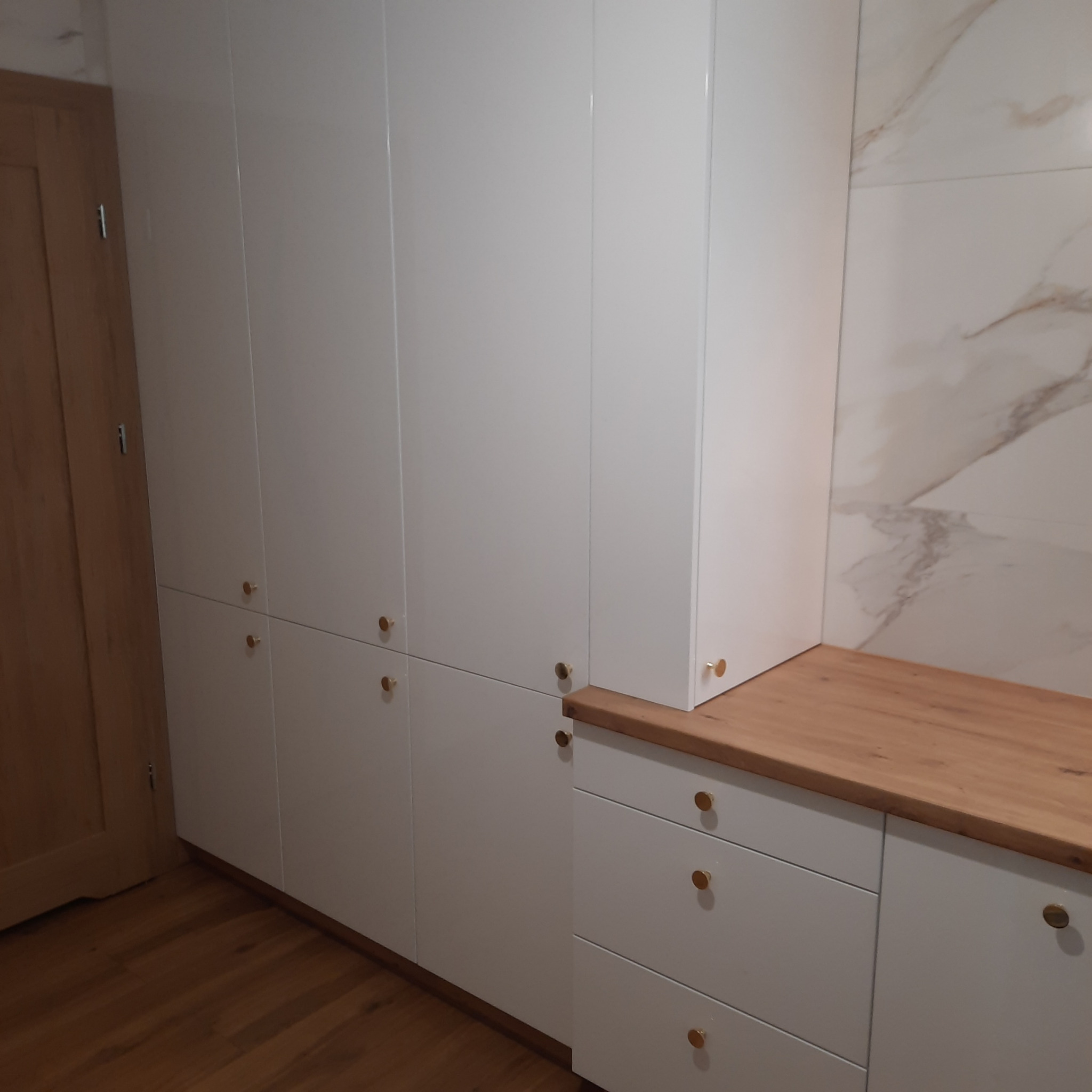 Biała zabudowa meblowa z drewnianym blatem i złotymi gałkami na tle marmurowej ściany, widoczny fragment drewnianych drzwi.