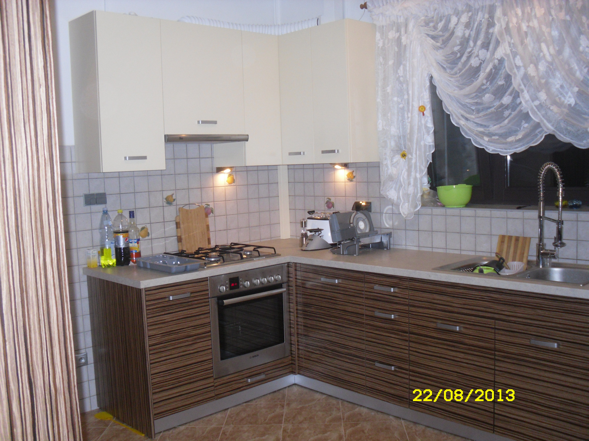 Jasna kuchnia narożna z frontami w poziome pasy, z piekarnikiem Bosch i płytą gazową, oświetlona halogenami podszafkowymi, z widocznymi butelkami i drobnymi akcesoriami na blacie.