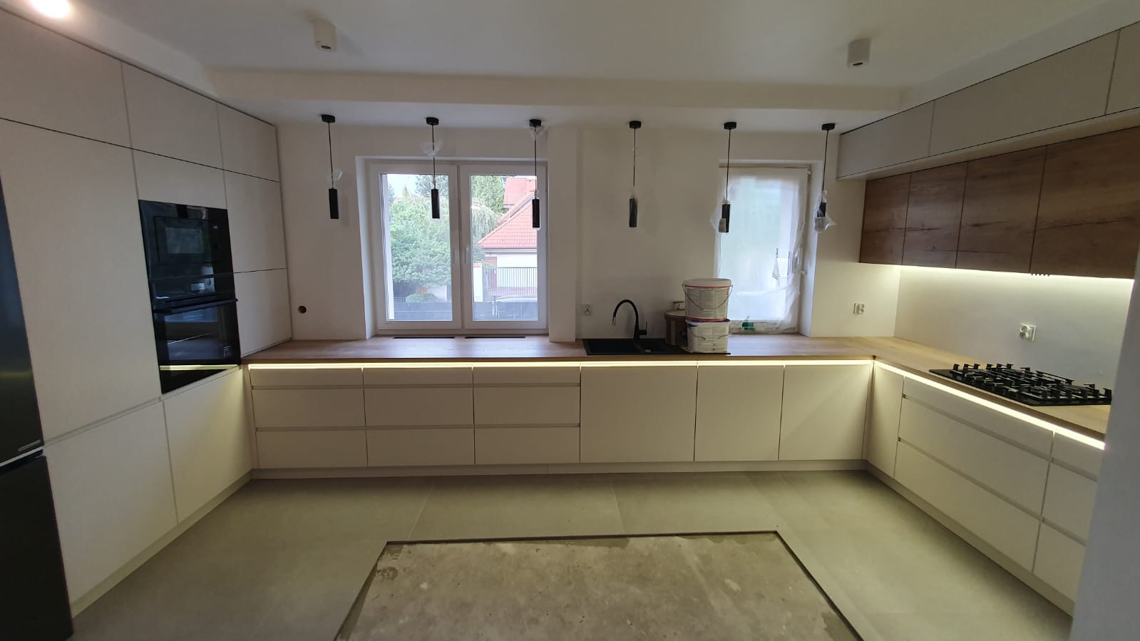 Nowoczesna kuchnia z białymi frontami, drewnianym blatem i podświetleniem LED pod szafkami, widoczne dwa wiadra z farbą przy oknie oraz fragment niedokończonej podłogi.