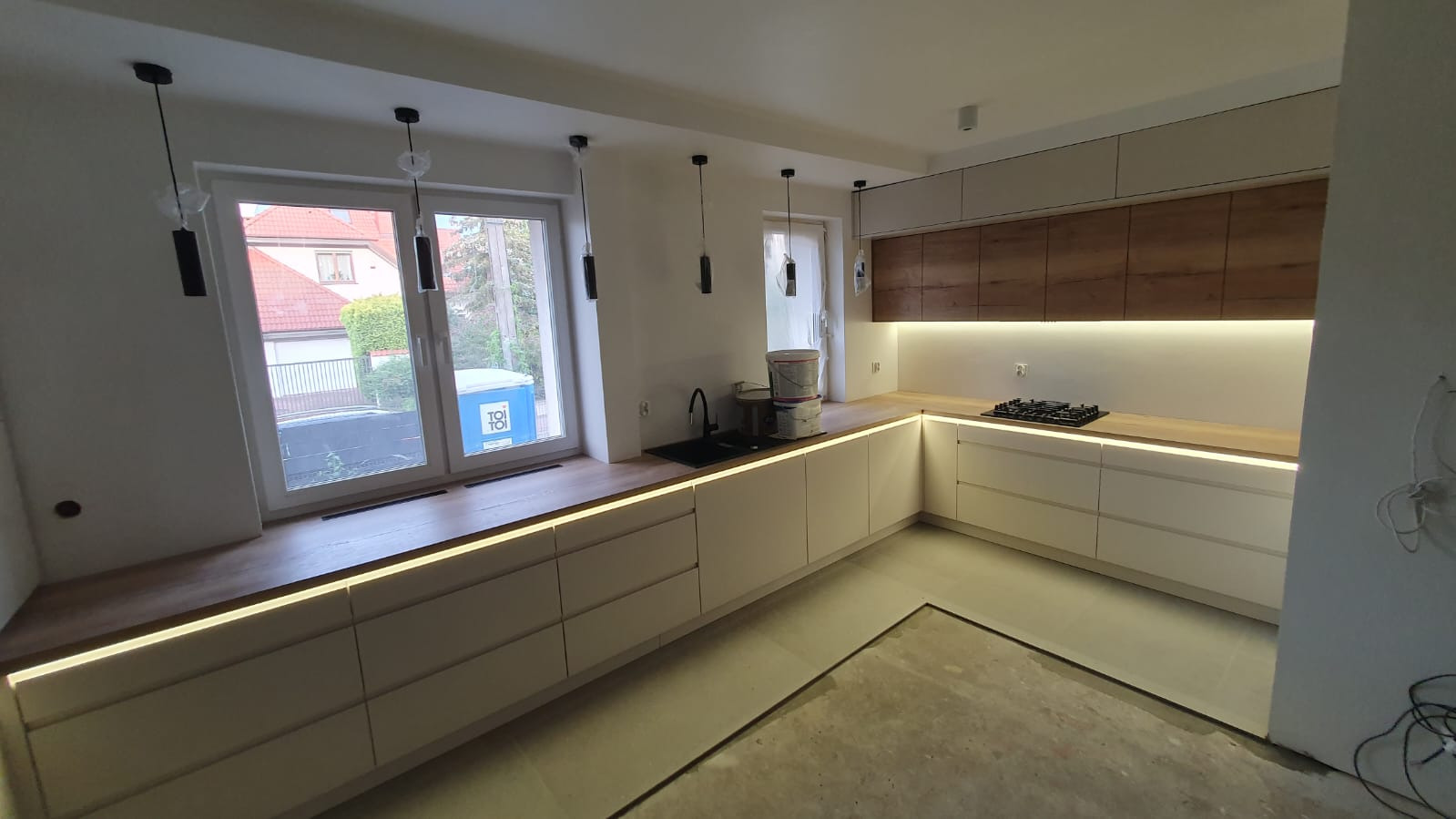 Nowoczesna kuchnia z białymi frontami i drewnianym blatem, podświetlana listwami LED, widok na zewnątrz przez okno z widocznym Toi Toi.