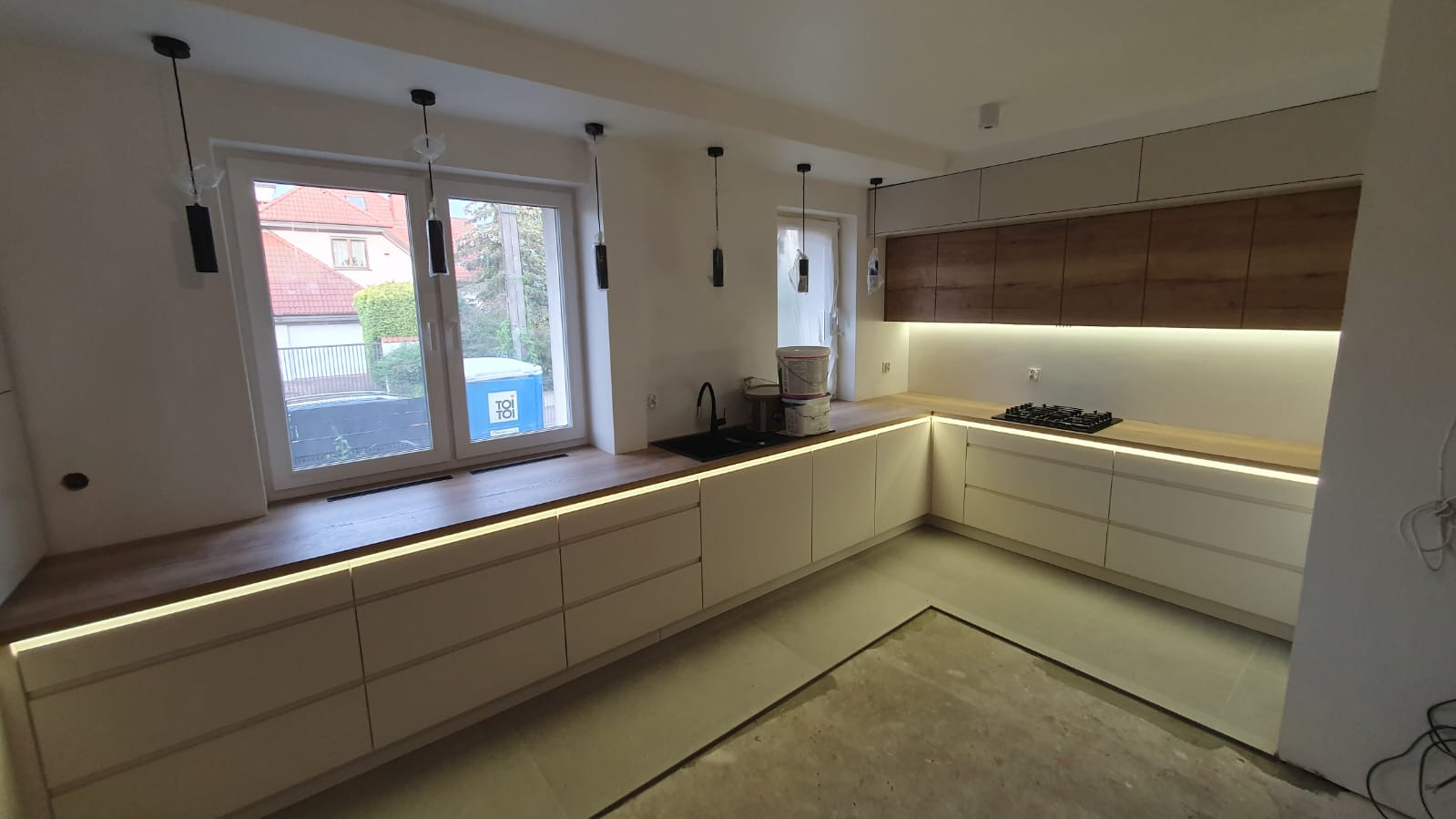 Nowoczesna kuchnia z białymi frontami szafek, drewnianym blatem i oświetleniem LED pod szafkami, widok na okno z pojemnikiem Toi Toi na parapecie, czarne lampy wiszące i niezakończona podłoga.