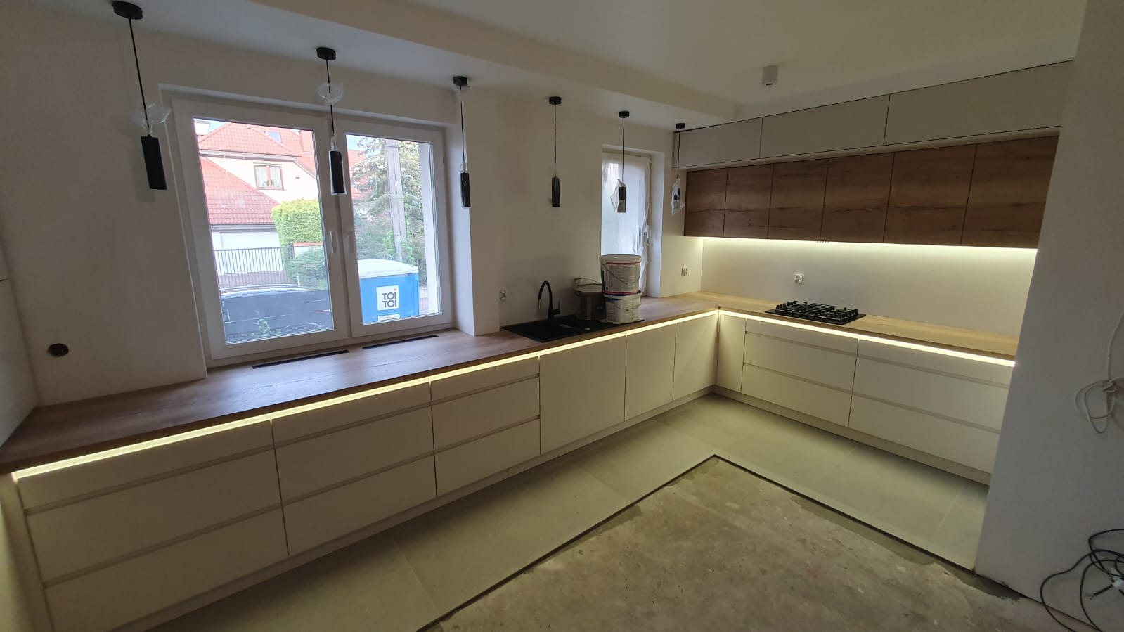 Jasna kuchnia na wymiar z drewnianymi blatami i podświetleniem LED, widoczne farby w wiadrach na blacie, niedokończona podłoga.