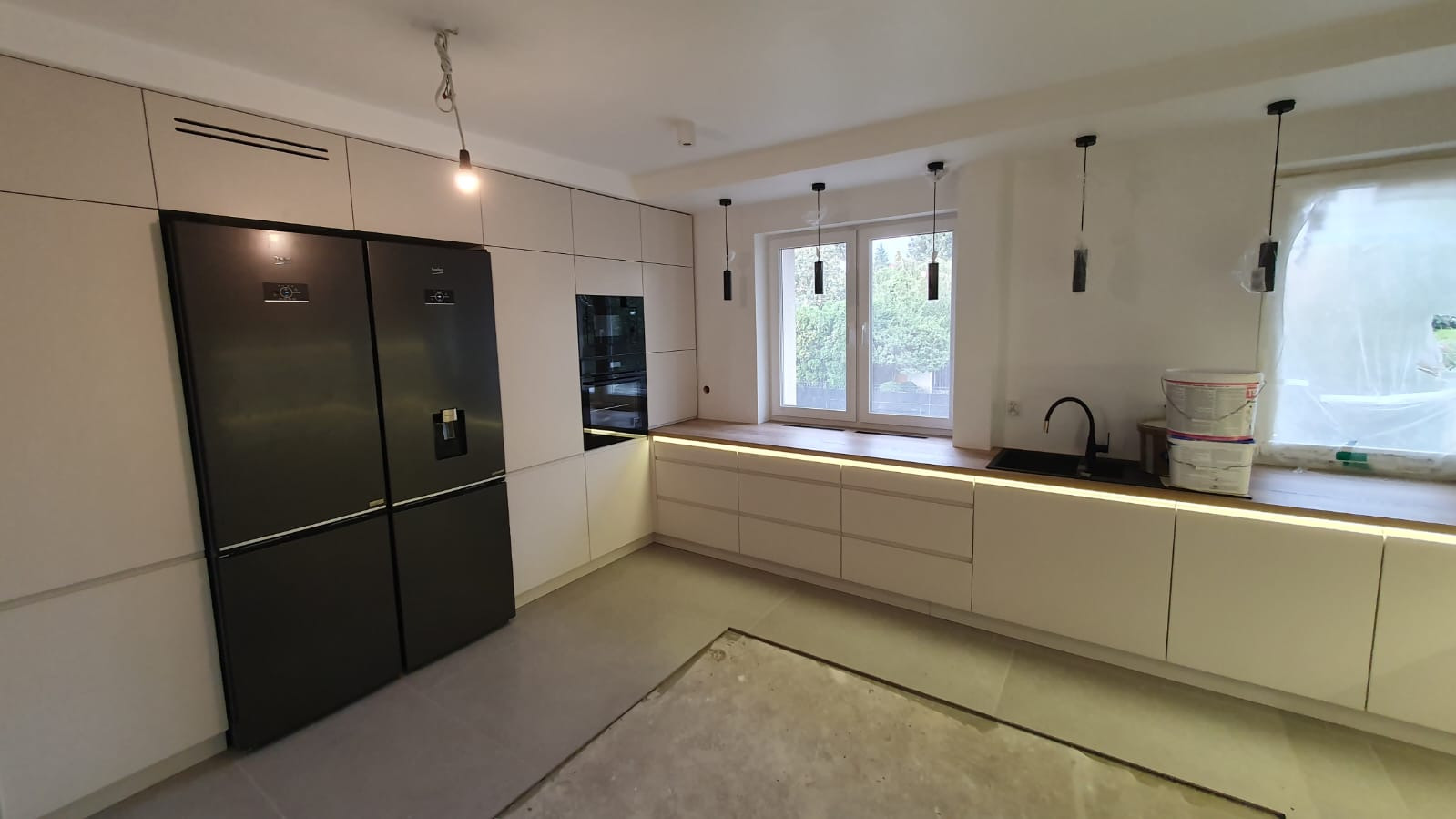 Nowoczesna kuchnia z białymi frontami, czarną lodówką side-by-side i oświetleniem LED pod blatem, widoczne puszki farby i niezakończone prace wykończeniowe na podłodze.