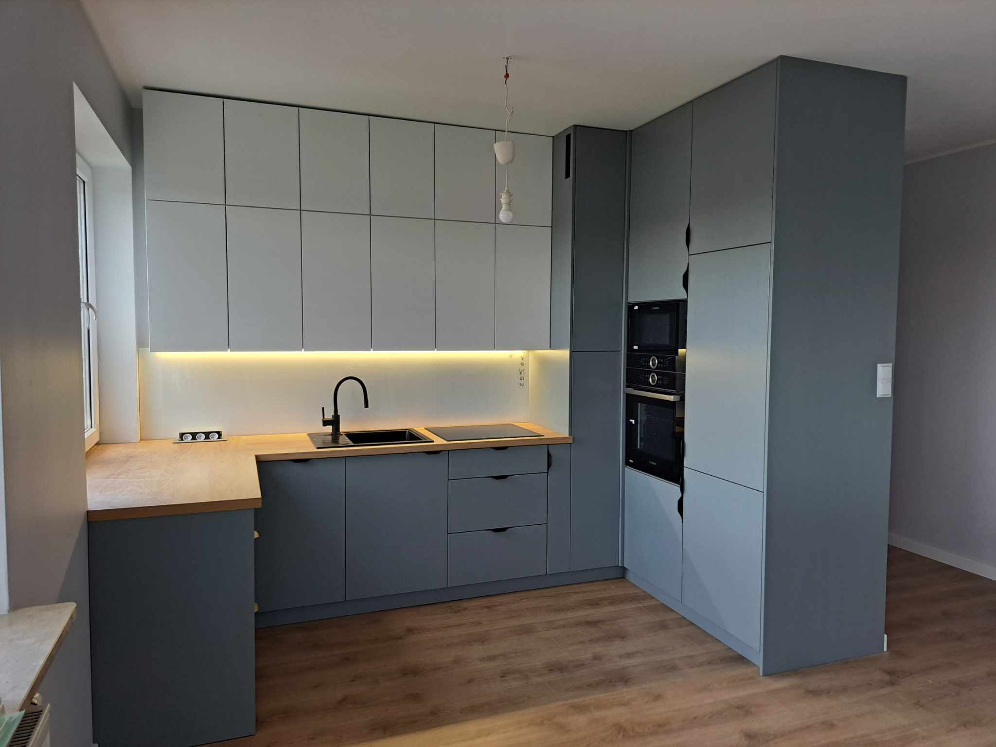 Nowoczesna kuchnia w odcieniach szarości z drewnianym blatem, oświetleniem LED pod szafkami wiszącymi i czarną armaturą, widok na zabudowę z piekarnikiem i mikrofalówką.