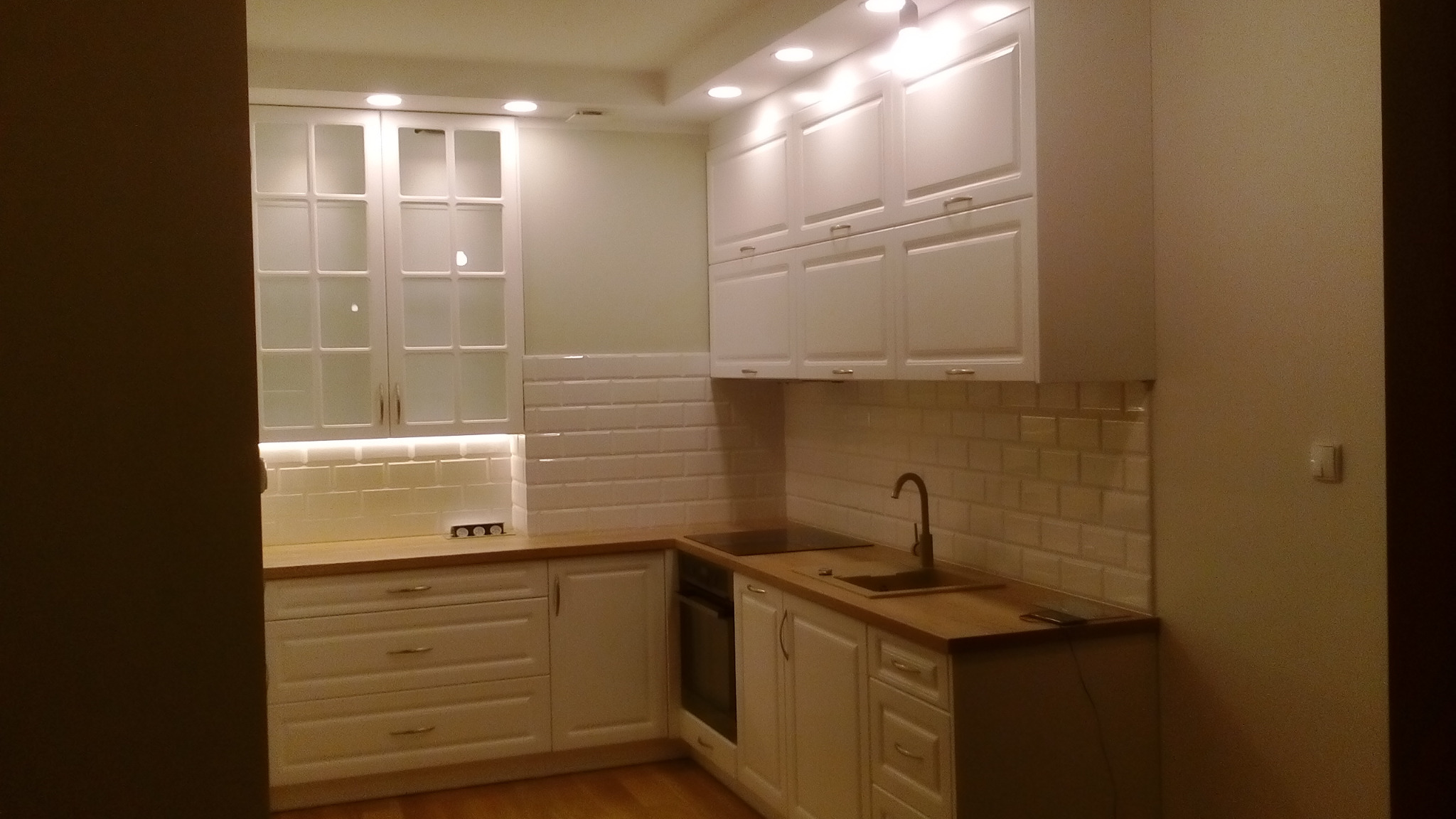 Jasna kuchnia z białymi frontami, drewnianym blatem i płytkami typu metro na ścianie, oświetlona punktowo i listwą LED pod szafkami wiszącymi.