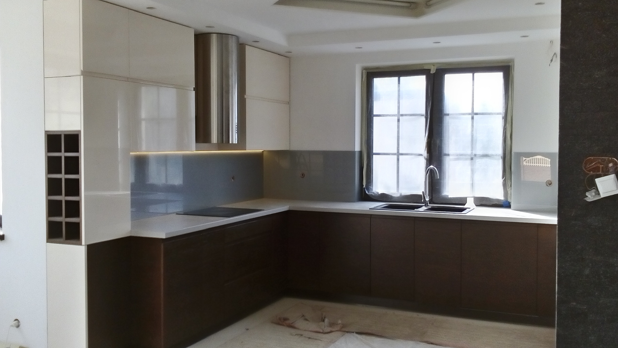 Nowoczesna kuchnia w trakcie montażu: białe i brązowe szafki, szary backsplash, stalowy okap, okno z zasłoniętą szybą, widoczne ślady po remoncie na podłodze.